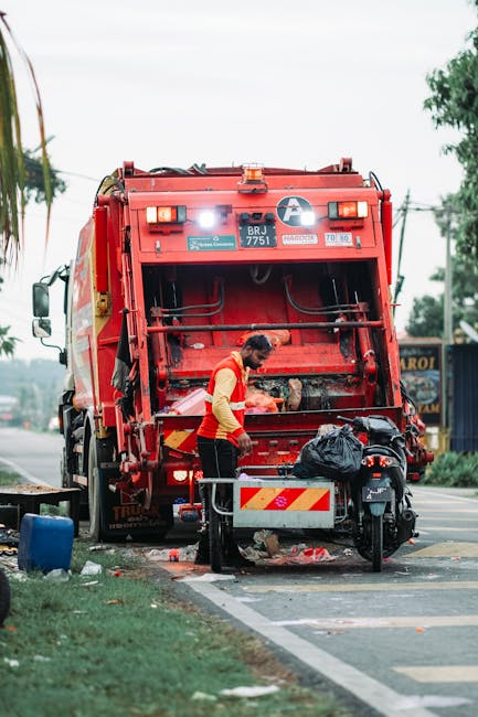 A worker wearing a yellow high-visibility vest and red shirt is seen operating a large red rubbish collection vehicle on a street. The truck has a prominent rear mechanism for compacting waste, with visible hydraulic parts and safety lights on top. The worker appears to be overseeing the collection process, possibly emptying a black refuse bag into the hopper. Nearby, a motorcycle is parked next to the truck, and a metal barrier with red and yellow reflective stripes is positioned at the back for safety. The surrounding environment includes a paved road with a curb, a grassy area, and some trees in the background, as well as a distant shop sign. Overhead, the sky is overcast, providing diffused natural light that softly illuminates the scene. This image illustrates the routine of on-site waste collection in an urban setting, highlighting how private rubbish removal services, such as those provided by Rubbish Removal Ealing, carry out efficient waste disposal without reliance on local authority services.