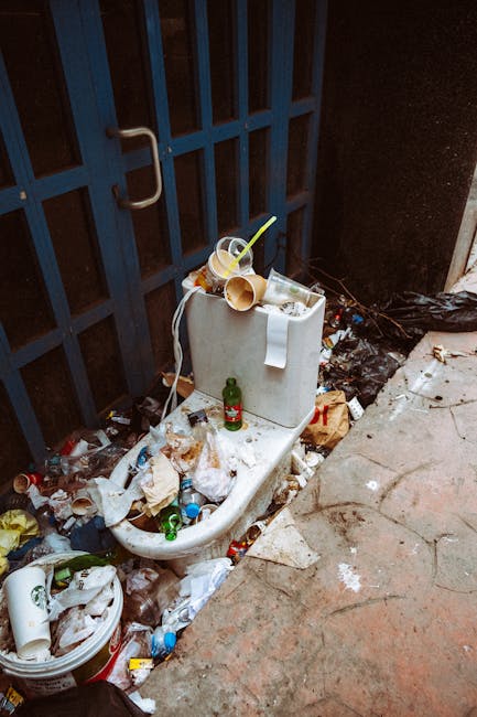 An overflowing toilet positioned against a dark metal gate in an outdoor area, surrounded by a large accumulation of assorted waste materials. The toilet bowl is urban-style ceramic, stained and partially obscured by discarded rubbish, including plastic bottles, paper, cardboard, and food packaging. Several crumpled paper towels and a cardboard paper towel roll are draped over the toilet tank, which is topped with used disposable coffee cups, a green glass bottle, and stray plastic straws. The ground around the toilet is littered with additional waste, including plastic containers, broken packaging, and crumpled wrappers, extending onto a weathered concrete surface and sidewalk. The environment appears neglected, with dirt and grime visible on the ground, indicating the waste has accumulated over time. This scene, captured in natural lighting, exemplifies an instance of private waste buildup that might be addressed through independent rubbish removal services by companies such as Rubbish Removal Ealing, specializing in on-site clearance and waste disposal solutions in urban outdoor settings.