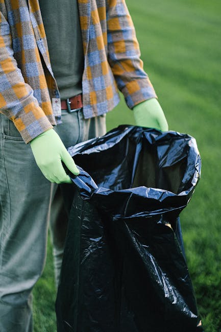 A person wearing a plaid shirt with shades of yellow, blue, and gray, along with gray trousers, is seen holding open a large black rubbish bag lined with a shiny black plastic interior. The individual is wearing bright green gloves and is standing outdoors on a grassy area, likely in a park or garden setting. The background features well-maintained green grass, indicating an environment suitable for gardening or outdoor waste collection. The scene captures the act of preparing or collecting waste materials, aligned with private rubbish removal or on-site clearance services provided by companies like Rubbish Removal Ealing, who handle various waste disposal needs away from local authority services. The image emphasizes cleanliness and manual handling of waste in an outdoor context, highlighting methods of independent collection or disposal of rubbish, possibly after tidying up a garden or outdoor space.