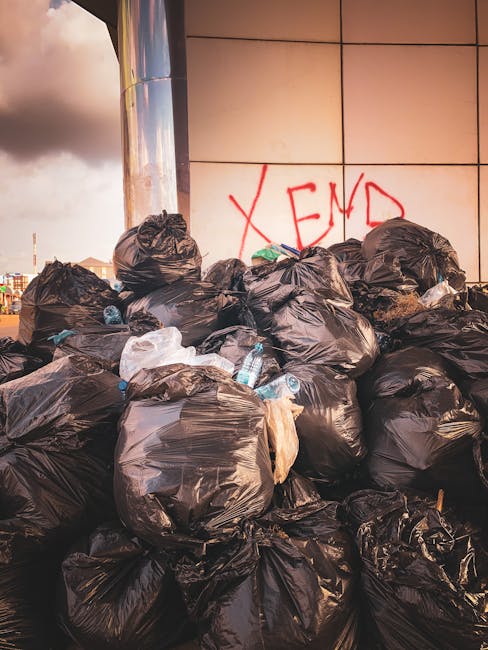 A large pile of black rubbish bags, some partially torn or spilling contents, stacked against a tiled wall with a red spray-painted message reading 'XEND' in an urban outdoor setting. Among the bags, several discarded plastic water bottles and smaller debris are visible. The environment appears to be in a city area, with a cloudy sky overhead, and the bags occupy a significant portion of the foreground, indicating an uncollected waste accumulation. The scene reflects an instance of excess waste that could require alternative rubbish collection methods, such as private disposal or on-site clearance services. The presence of the bags in front of a modern tiled building suggests a commercial or retail area that might benefit from professional rubbish removal, aligning with services offered by Rubbish Removal Ealing.
