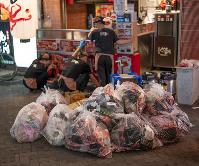 Several large transparent plastic bags filled with mixed waste, including clothing and possibly other recyclables, are placed on the pavement outside a shop or fast-food outlet. The bags are piled together, some appearing loosely closed with visible contents, in front of a group of three workers wearing black uniforms with logo patches, who are crouched down or standing near the store's entrance. The background shows a brick wall, a vending machine or waste disposal unit, and various signs and boxes, indicating an urban setting. The scene is illuminated by outdoor lighting, casting soft shadows on the ground. This image exemplifies on-site waste collection and preparation for rubbish removal, consistent with private waste management services such as those offered by Rubbish Removal Ealing, highlighting the process of gathering and handling rubbish in commercial or communal areas for efficient collection and disposal.