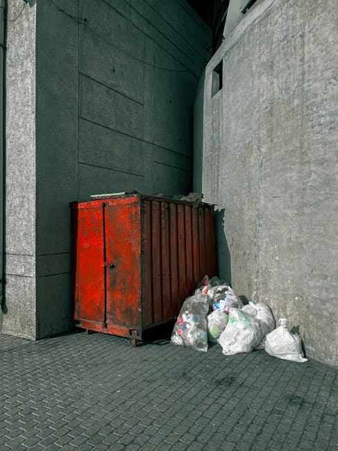 A large, weathered red metal skip with rust spots and a slightly dented surface is positioned against a dark green wall on the left and a light grey concrete wall on the right, in a narrow urban alleyway. The skip, used for waste collection, is filled with assorted rubbish that is spilling out onto the paved ground. Surrounding the skip are multiple white plastic bags filled with discarded waste, some tightly knotted and others loosely closed, resting on the textured grey paving stones. The scene is illuminated by natural daylight, casting minimal shadows, and the environment appears clean but utilitarian, typical of private waste disposal efforts outside a commercial or residential building. This setup by Rubbish Removal Ealing exemplifies an on-site rubbish clearance scenario, highlighting the type of waste typically handled during independent collection services for timely rubbish removal in the London area.